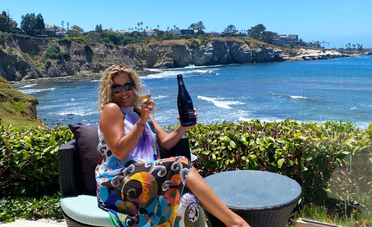 Nina Snow, owner of DOCG Imports, holding a bottle of Italian wine with La Jolla Cove in the background.